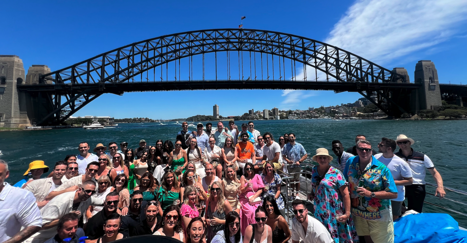 A large group of people, including representatives from Lending Association, pose for a photo on a boat with the Sydney Harbour Bridge in the background under a clear blue sky.